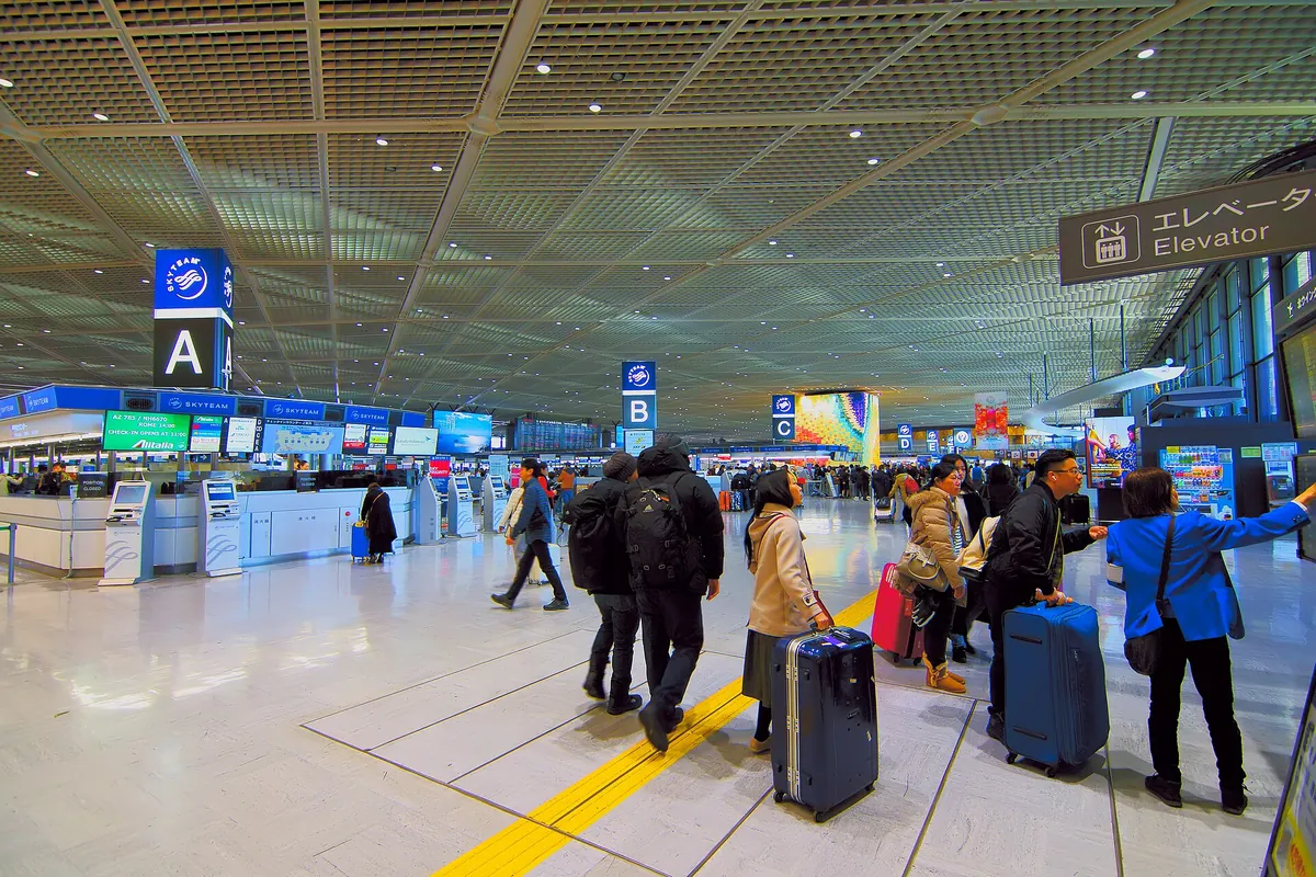 成田空港の出発ロビーでA、B、Cの看板が立つチェックインカウンターの前をスーツケースを引いた旅行者が歩いている様子