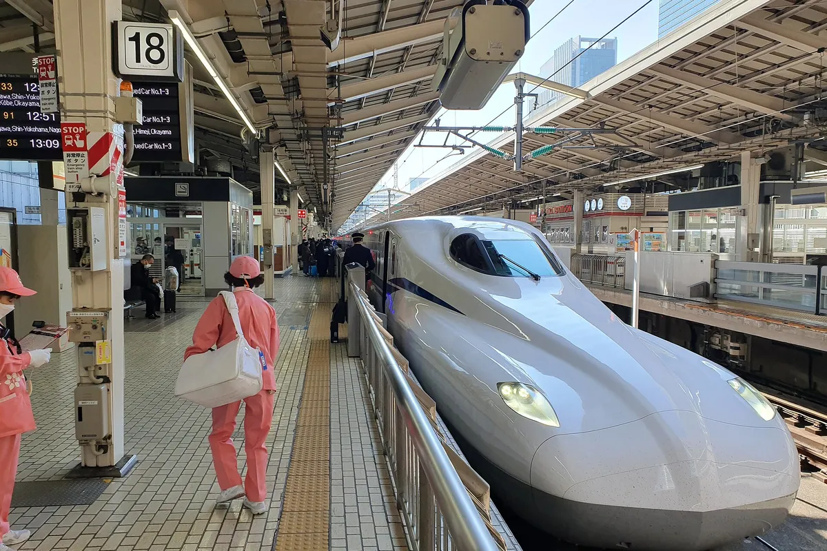A white Shinkansen bullet train standing at platform 18, with a person in a pink uniform walking along the platform