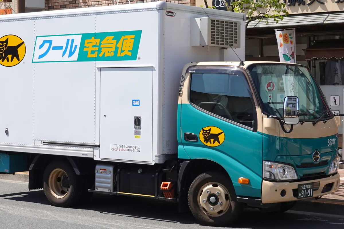 A teal and white Yamato Transport refrigerated delivery truck with a black cat logo parked in front of shops in Japan