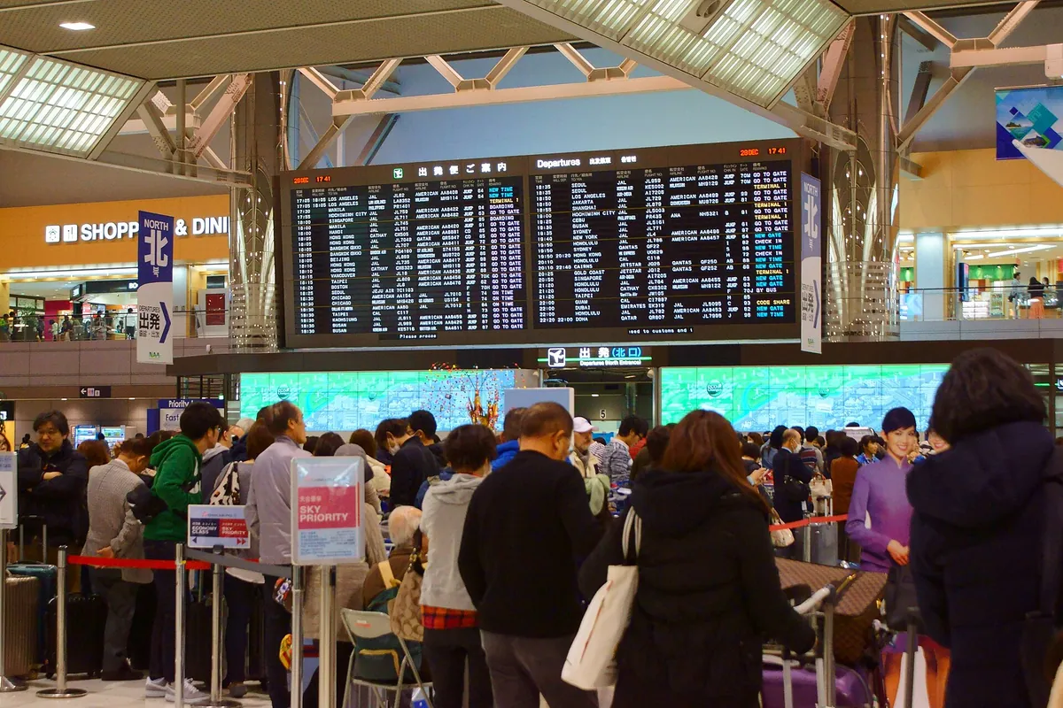 A crowded departure lobby at Narita Airport Terminal 2 with travelers and suitcases queuing beneath a flight information boar