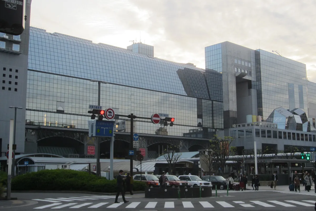 The glass facade of Kyoto Station's main building with taxis and pedestrians crossing the street in front