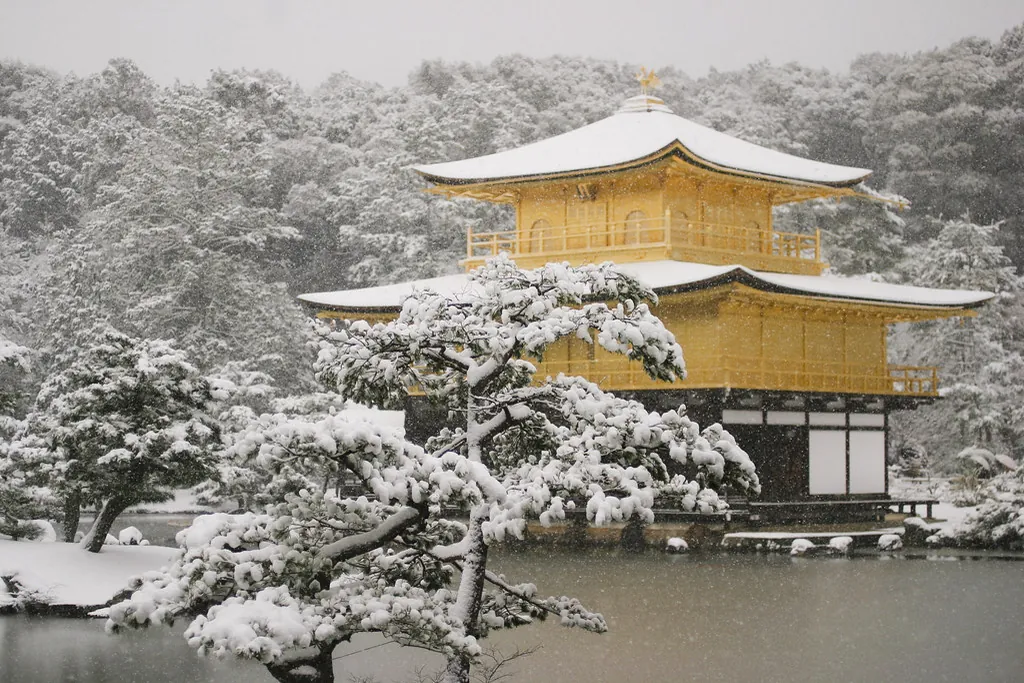 Kinkaku-ji golden pavilion in Kyoto with snow covering its roof and pine trees, snow falling over the pond
