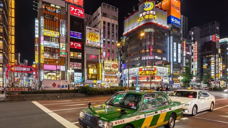 A green and yellow taxi at a nighttime intersection in Shinjuku, Tokyo, with illuminated signboards and a white taxi nearby.