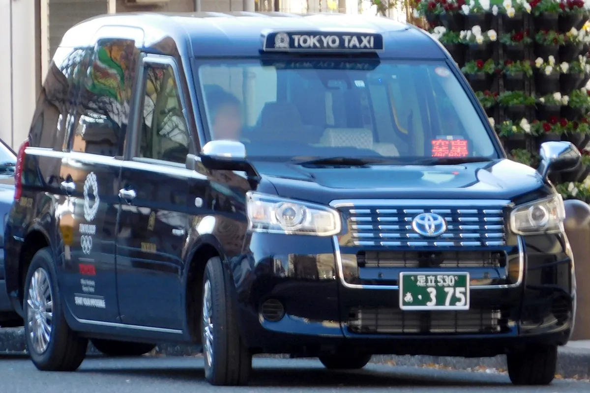 Front view of a black Toyota JPN Taxi with an illuminated TOKYO TAXI roof sign, parked beside flower displays.