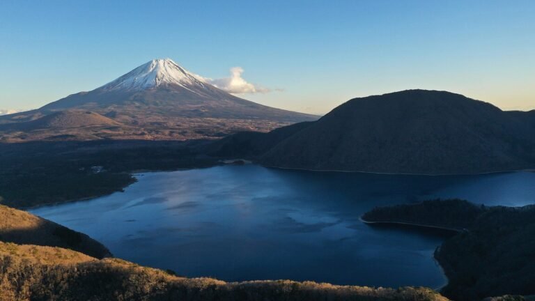 雪をかぶった富士山が湖の上にそびえ、周りに山や丘が広がる日本の風景