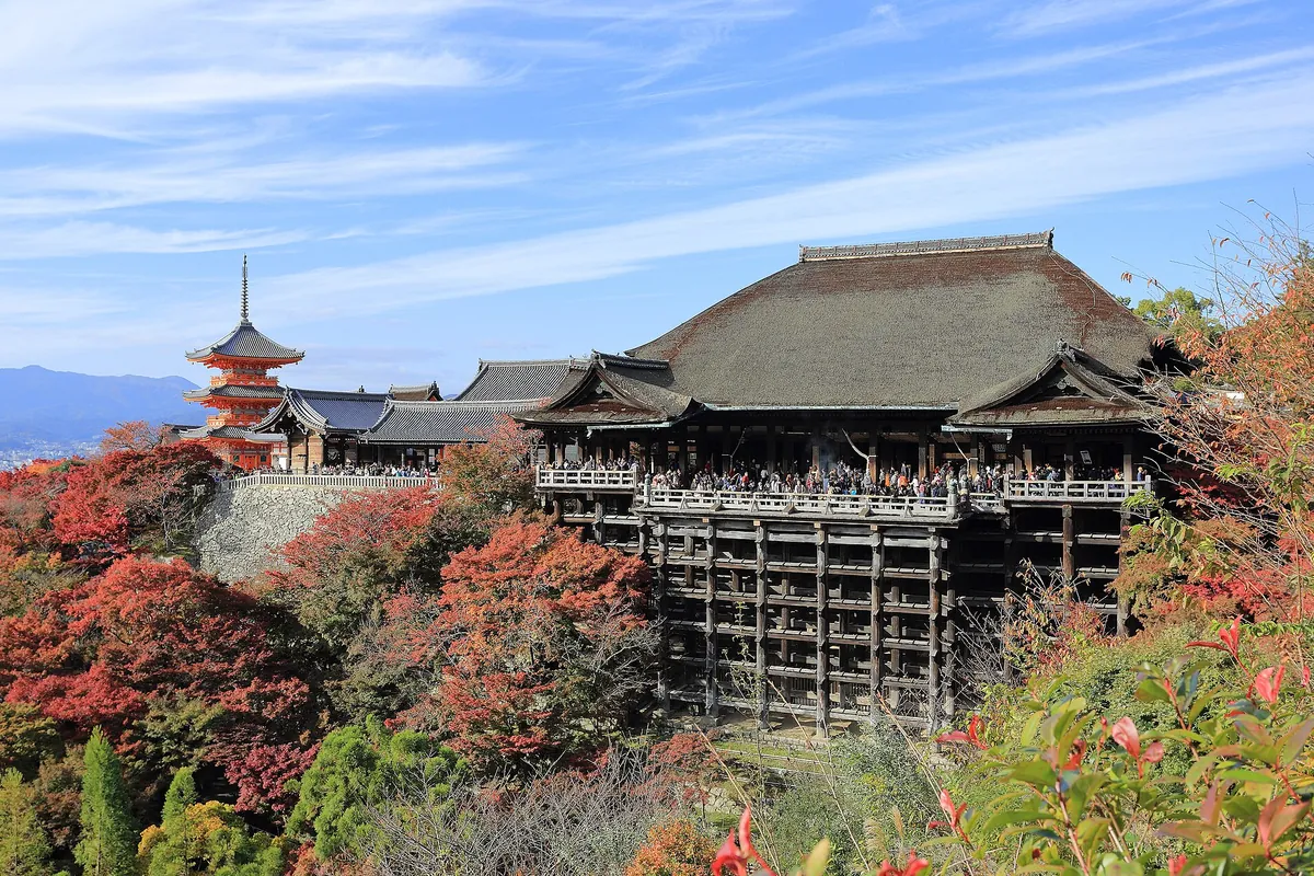 The wooden stage of Kiyomizu-dera temple built into a hillside, with crowds of visitors and autumn foliage in Kyoto