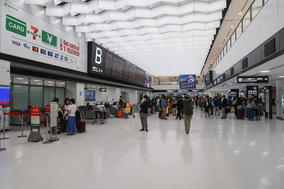 Arrivals hall at Narita Airport Terminal 2 with travelers walking past signs and a currency exchange counter