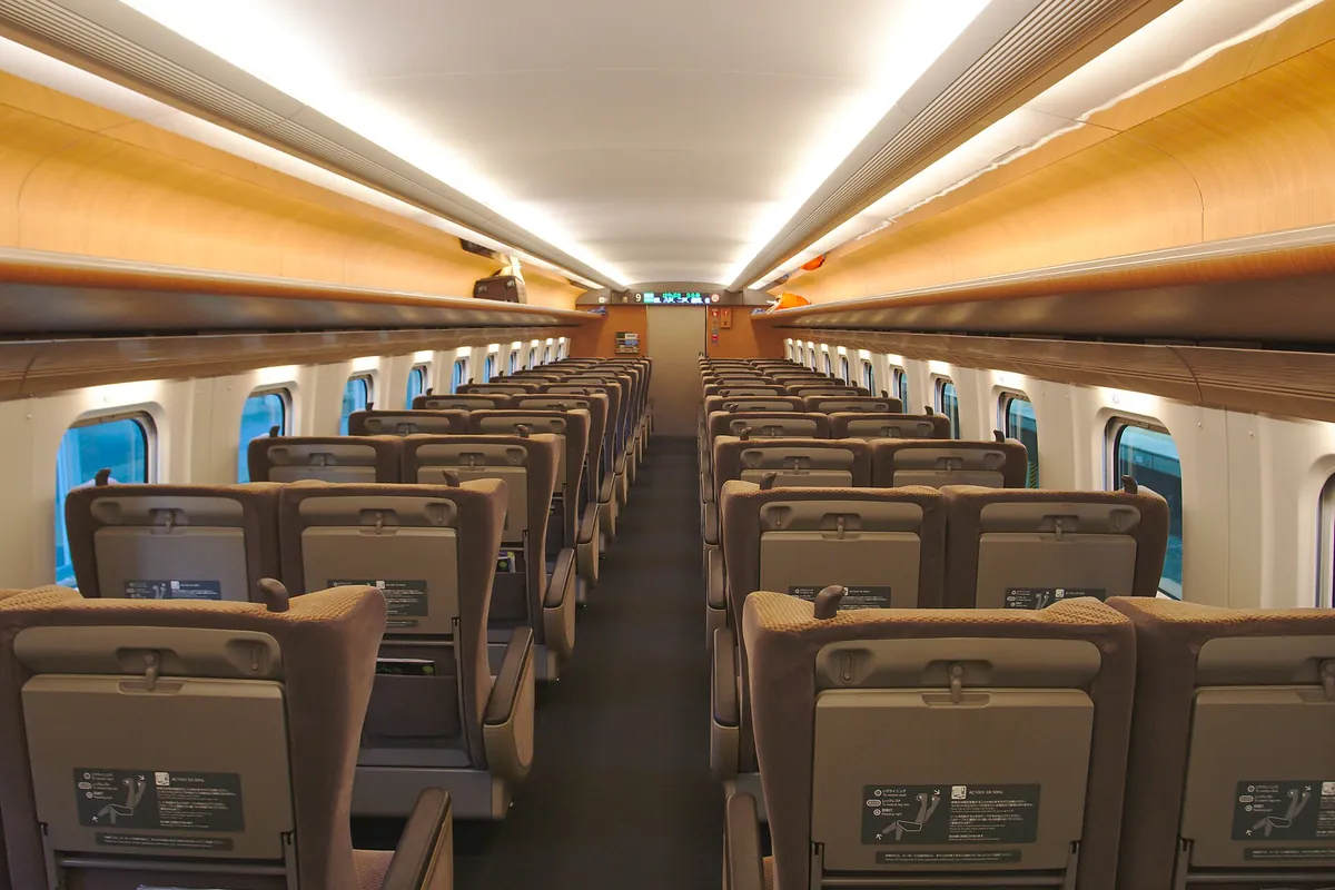 Rows of tan Shinkansen seats with headrests lining a center aisle under a wood-paneled ceiling