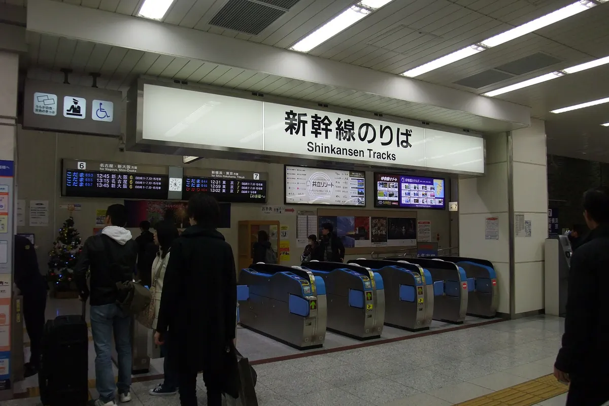 Automatic ticket gates below a Shinkansen Tracks sign with a departure board, a small Christmas tree and passengers nearby