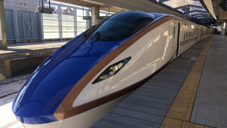 White E7 series Hokuriku Shinkansen train with a blue nose and copper stripe at a snowy station platform