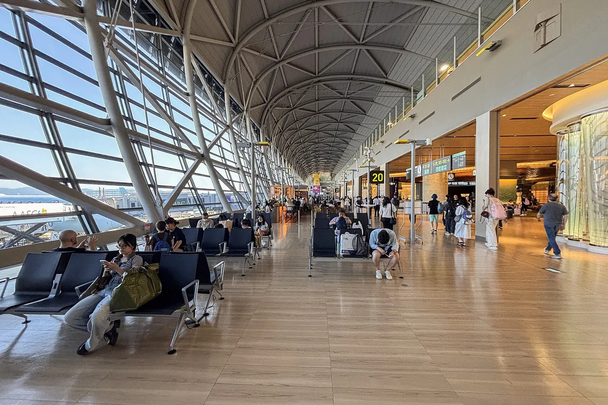 Terminal corridor at Kansai Airport with a curved ribbed roof, rows of seating, a gate 20 sign, and travelers walking past