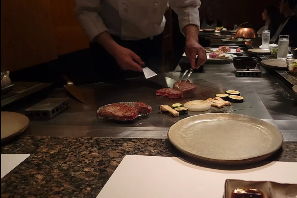 A chef grilling marbled Kobe beef and vegetables on a teppanyaki griddle