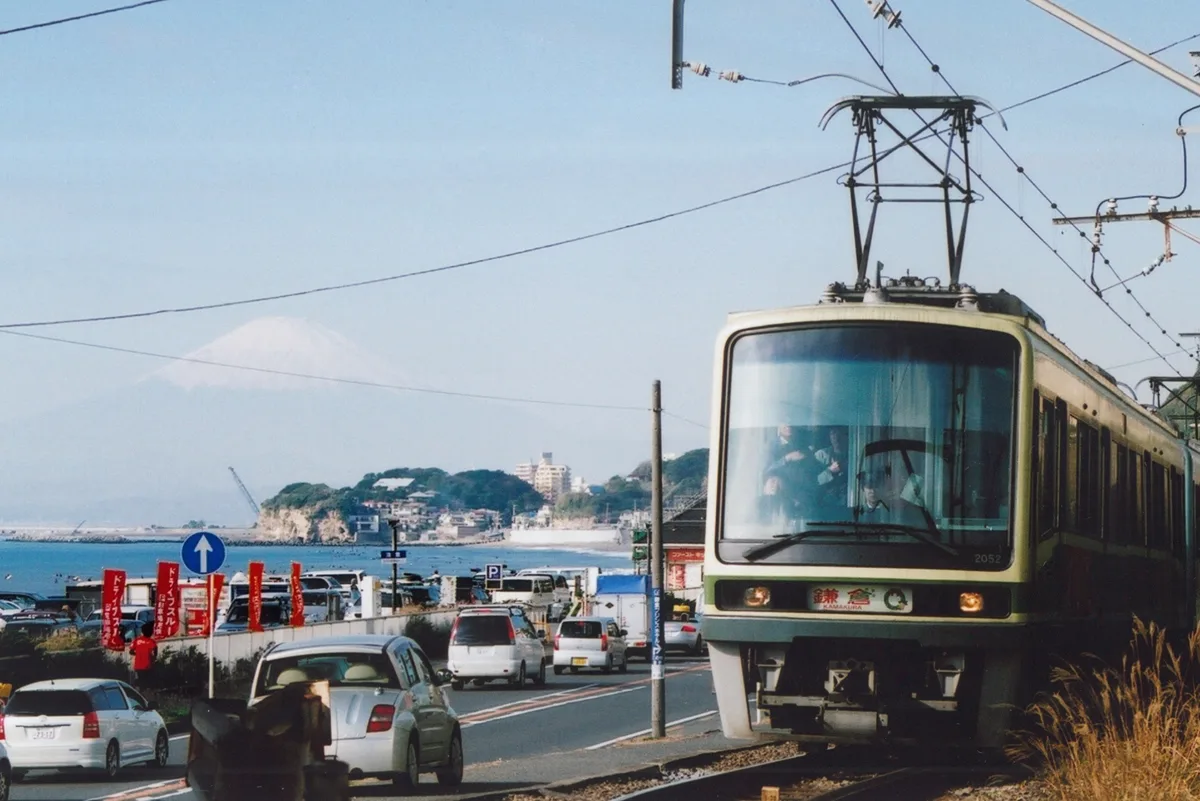 A green and cream Enoden train running beside the Shonan coast road, with the sea, Enoshima and Mount Fuji in the distance.