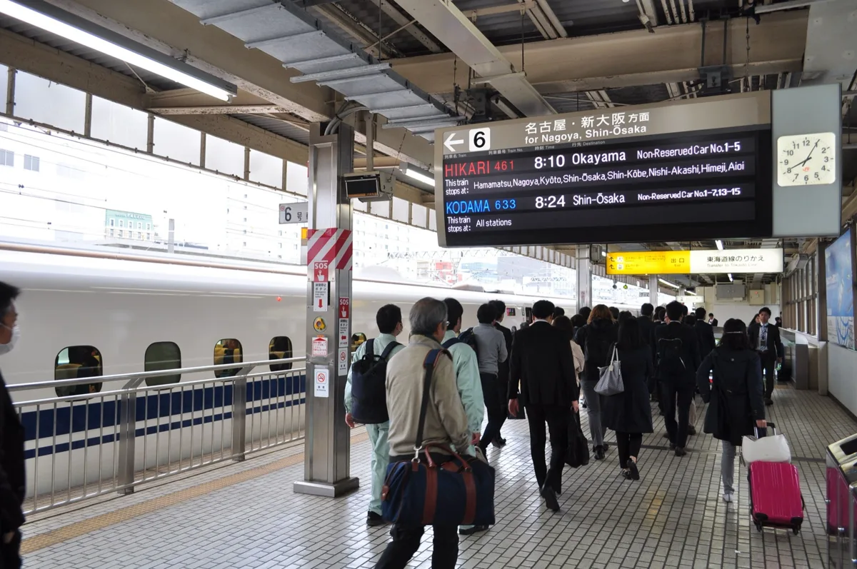 A departure board at Shizuoka Station showing a Hikari to Okayama and a Kodama to Shin-Osaka, with passengers walking on the.