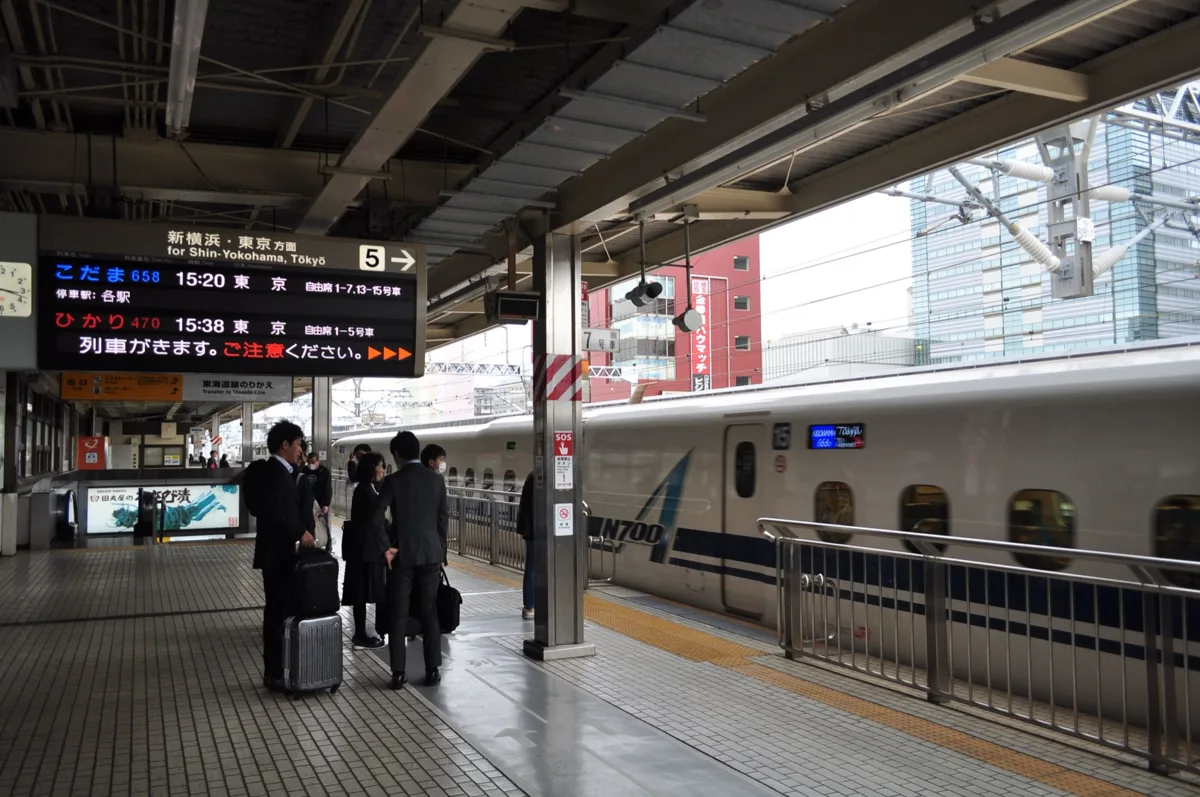 A departure board at Shizuoka Station showing a Kodama and a Hikari to Tokyo, with passengers waiting by an N700 series.
