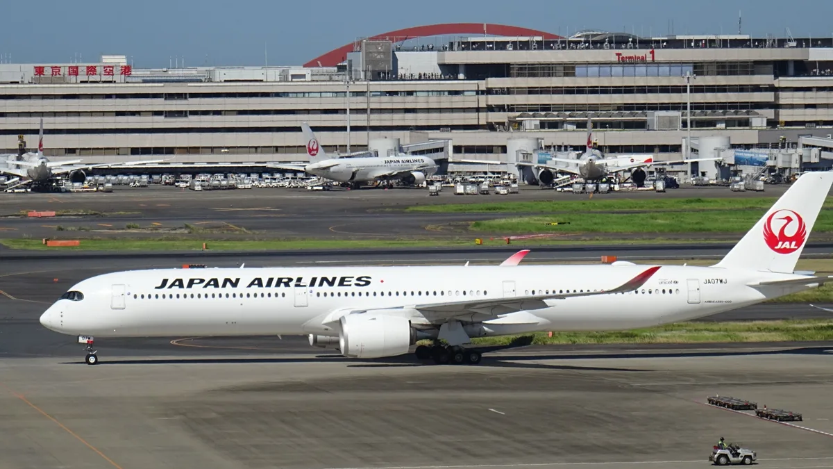A Japan Airlines Airbus A350-1000 taxiing in front of Haneda Airport Terminal 1, with the Tokyo International Airport sign.