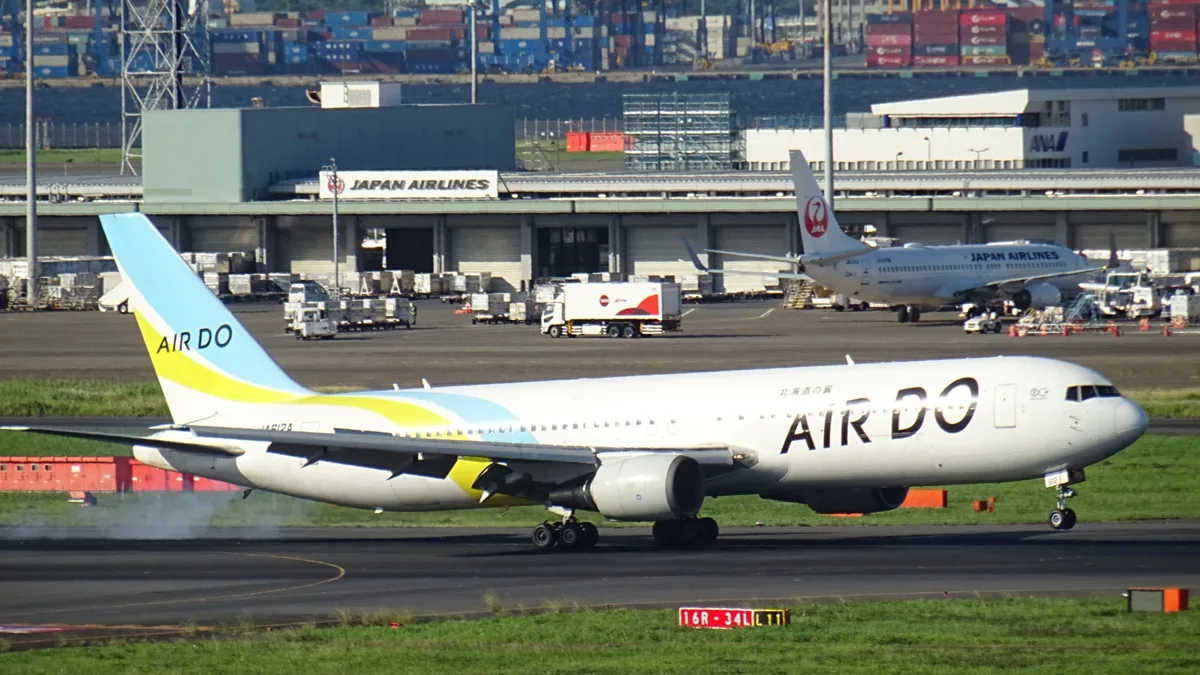 An AIRDO Boeing 767-300 with a light blue and yellow tail on the runway at Haneda Airport, with a JAL aircraft behind, in.