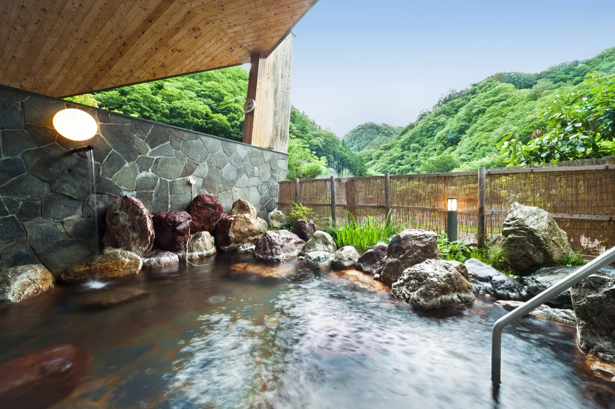 An empty outdoor hot spring bath (rotenburo) with rocks and a bamboo fence, with green hills behind, in 2020