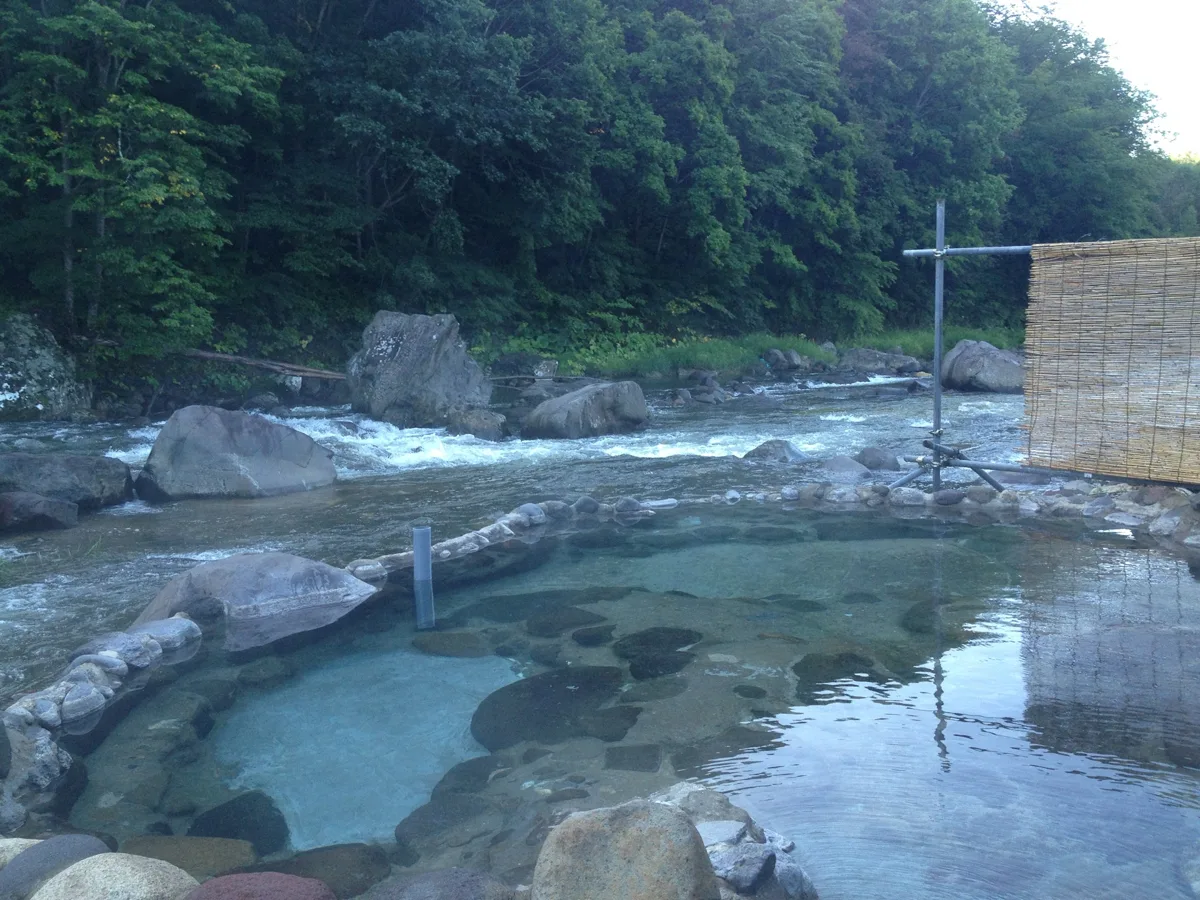An empty riverside outdoor hot spring bath (rotenburo) made of stones, right next to a fast-flowing mountain river, in 2018