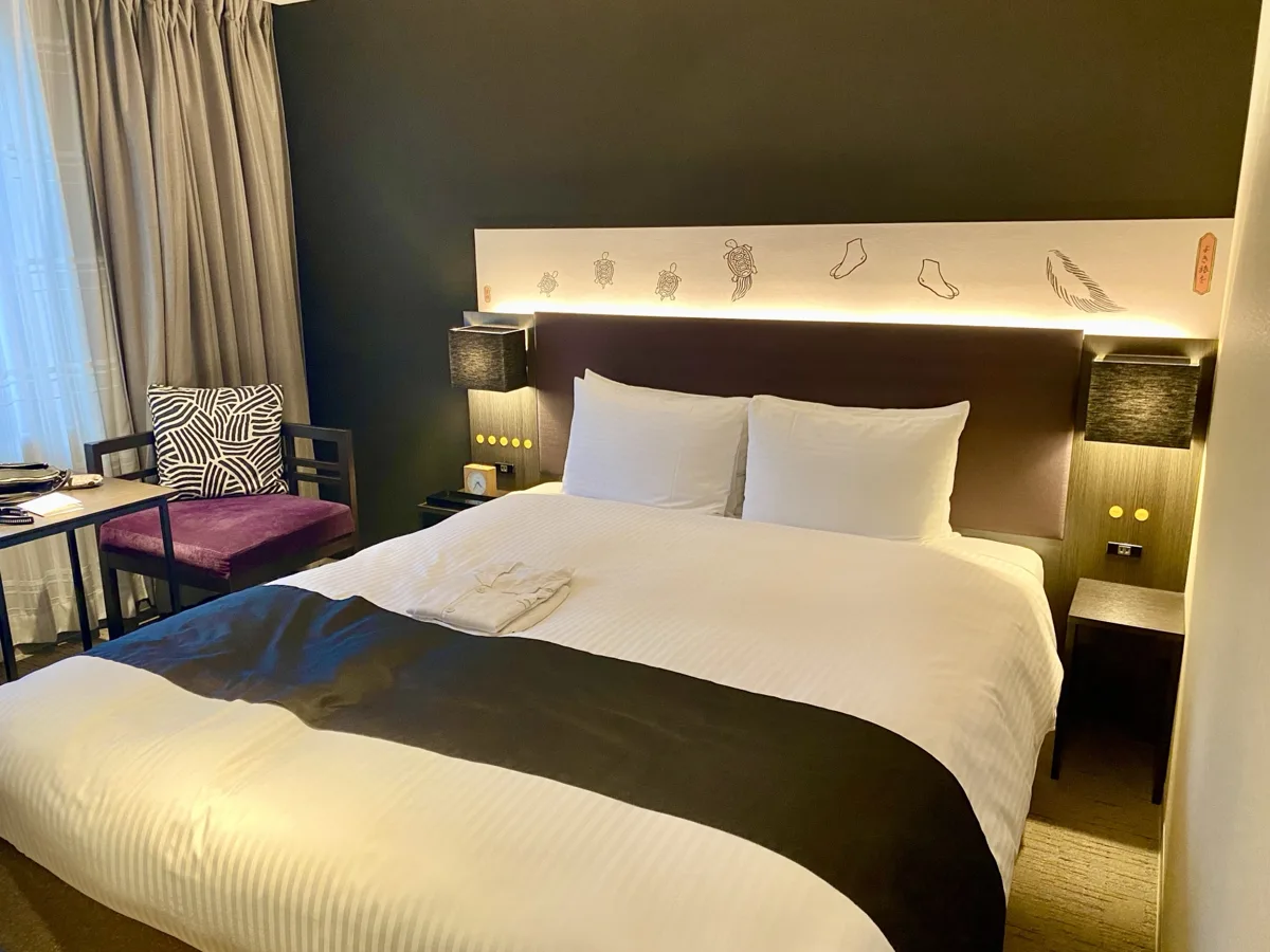 A modern hotel guest room in Tokyo with a large double bed, a dark bed runner and a small sofa by the window, in 2024
