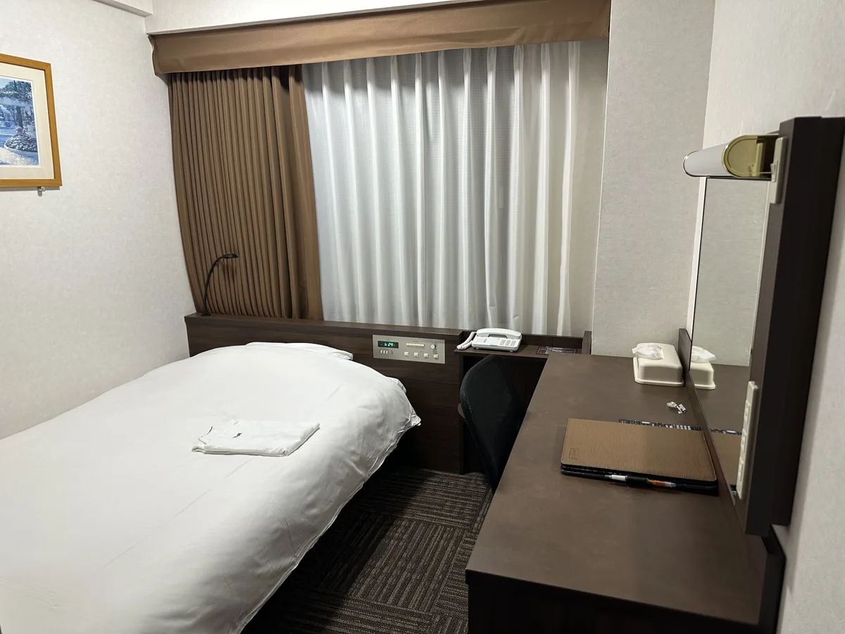 A small single room in a Japanese business hotel, with a narrow bed, a desk with a mirror and a curtained window, in 2025