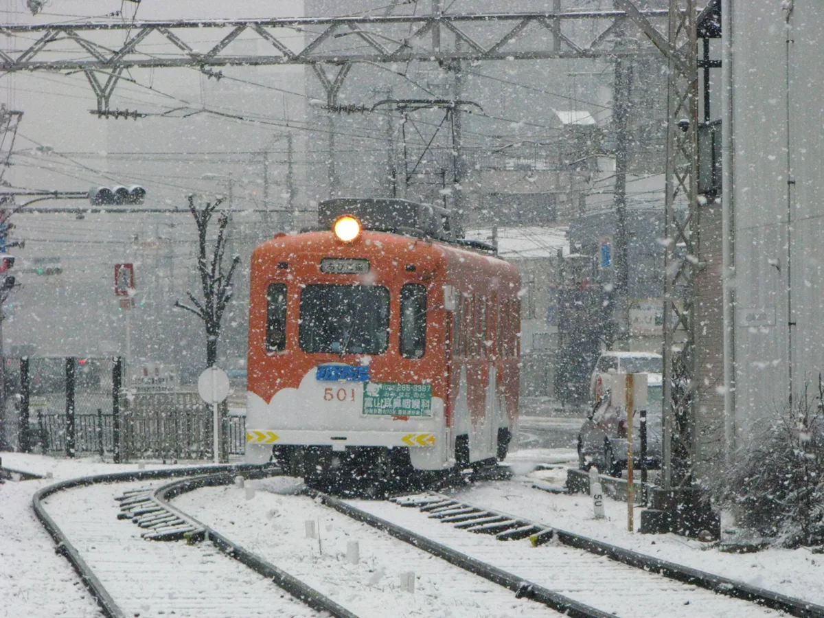 An orange Hankai Tramway streetcar running through falling snow on a snow-covered track in Osaka
