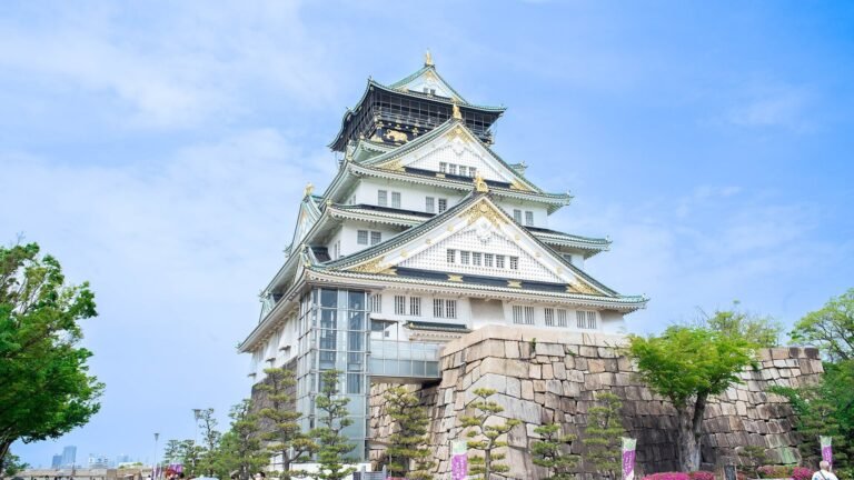 The white and green main keep of Osaka Castle with gold trim, seen from below with stone walls and trees