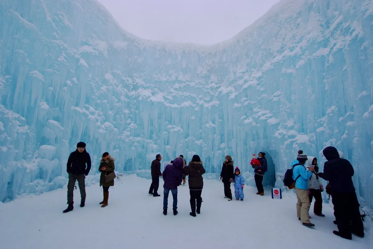 Pale blue ice walls forming a circular enclosure at the Lake Shikotsu Ice Festival, with visitors standing inside in daylight