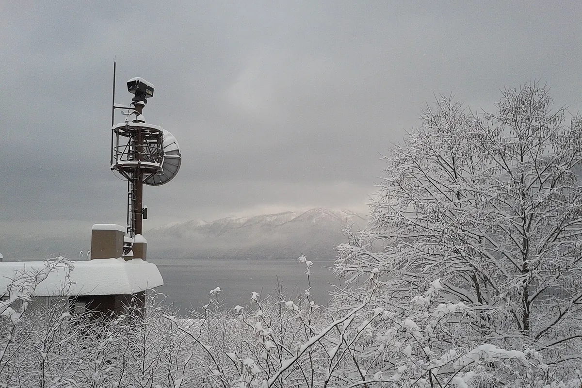 Lake Shikotsu in winter seen through snow-covered trees, with a radar tower and a snow-dusted mountain across the water