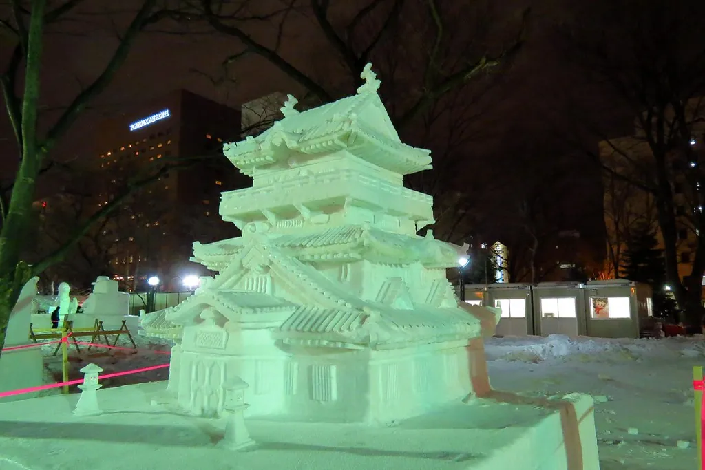 A large snow sculpture of a castle lit up in green at the Sapporo Snow Festival at night, surrounded by bare trees