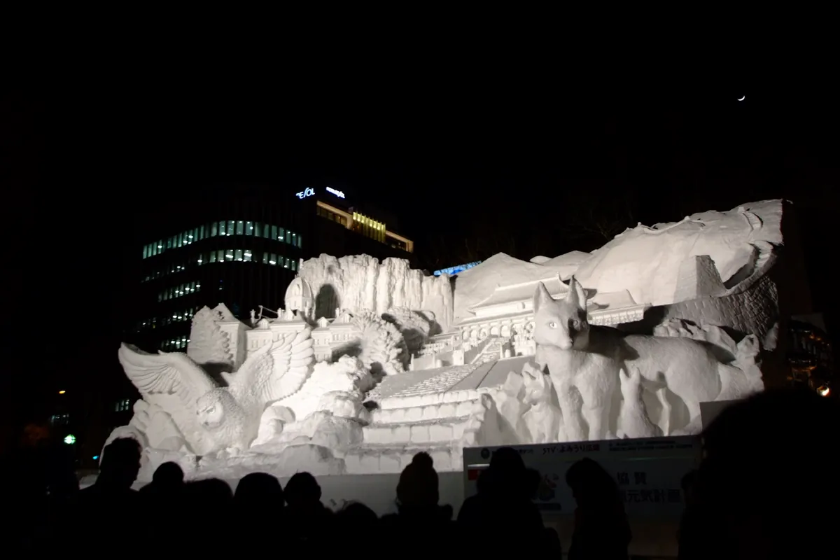 A large illuminated white snow sculpture at the Sapporo Snow Festival at night, with spectators watching