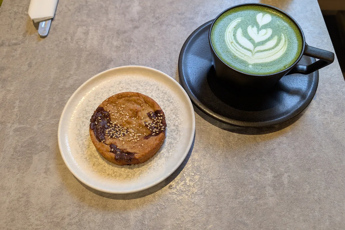 A matcha latte with rosetta latte art in a black cup on a saucer, beside a round cookie topped with sesame seeds on a plate