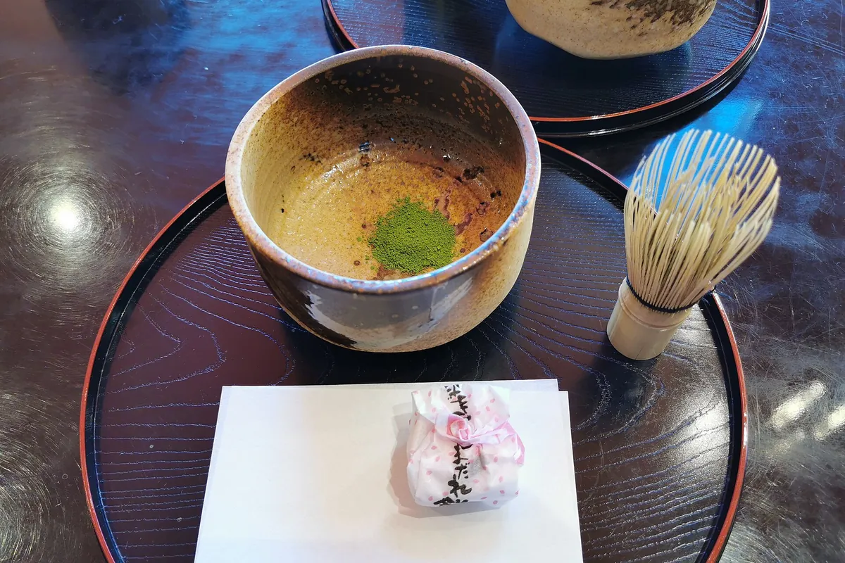 A ceramic bowl with a small heap of matcha powder and a bamboo whisk beside it, with a wrapped sweet on a napkin