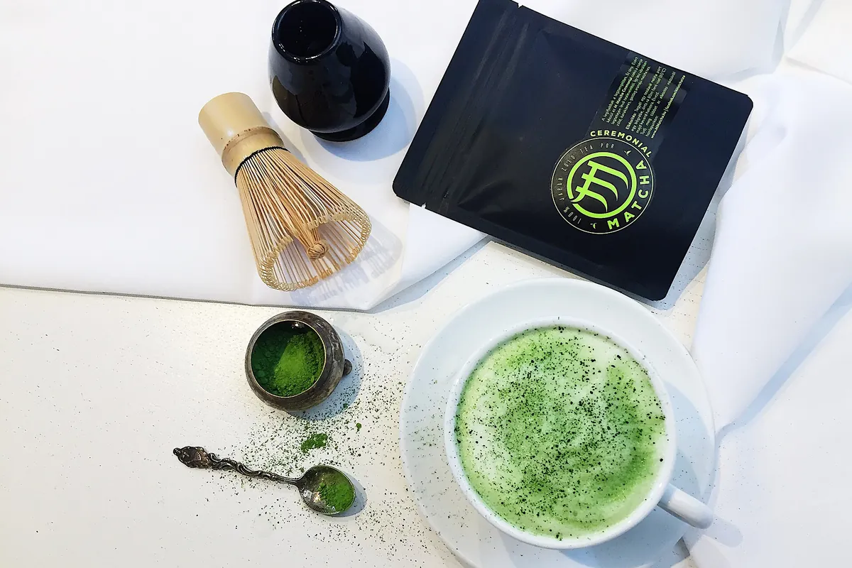 A bamboo whisk, a bag of matcha powder, a small bowl of matcha, a spoon, and a cup of matcha latte on a table