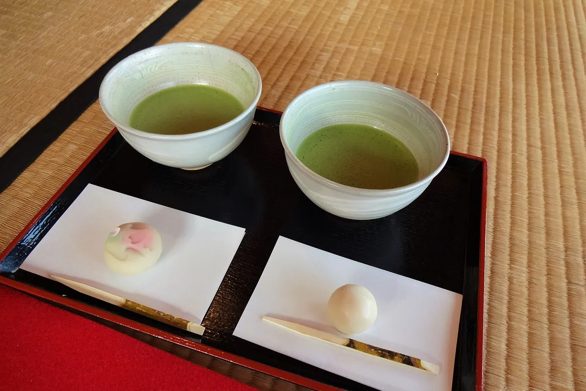 畳の床に置かれた黒いお盆に、抹茶が入った茶碗2つと和菓子2つが紙の上に並んでいる様子