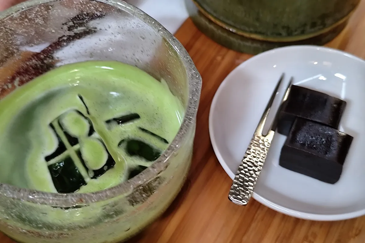 木製のテーブルに置かれたグラスカップ入りの抹茶クリームデザートと、濃い色の立方体2つとフォークが載った皿