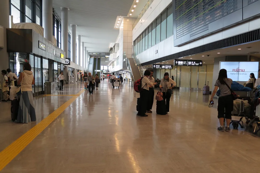 成田空港内の通路で、旅行者と荷物カート、フライト案内板、エスカレーターが見える様子