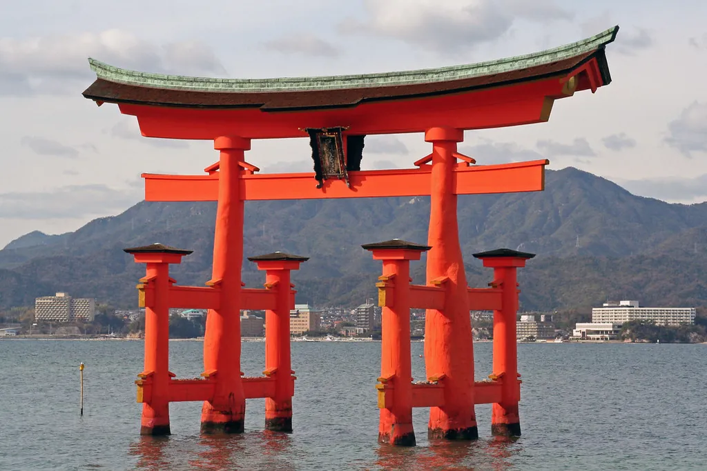 The vermilion torii gate of Itsukushima Shrine standing in the sea with mountains and buildings behind