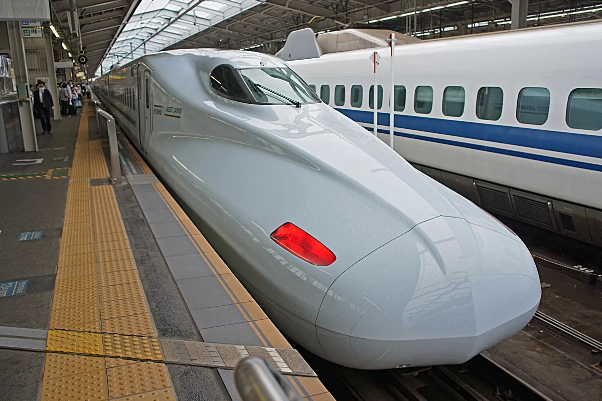 The pale blue nose of an N700 series Shinkansen train beside a blue-and-white Shinkansen train at a platform