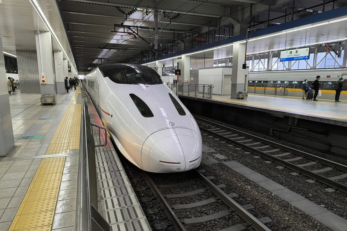 A white Kyushu Shinkansen 800 series train stopped at a platform at Hakata Station with passengers nearby