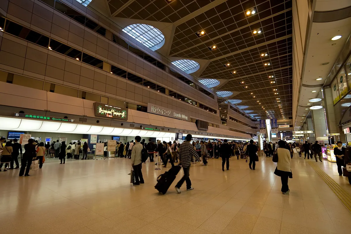 A wide airport departure hall with check-in counters, a security check sign, and travelers pulling luggage