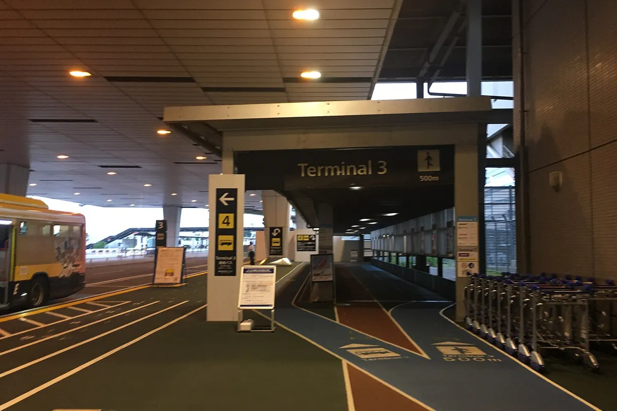 Narita Terminal 3 sign above a covered walkway, with blue and reddish-brown lanes on the floor and luggage carts