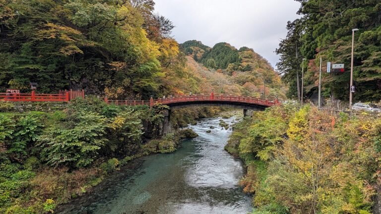 The red Shinkyo Bridge arching over the Daiya River in Nikko, with autumn-colored trees along the banks