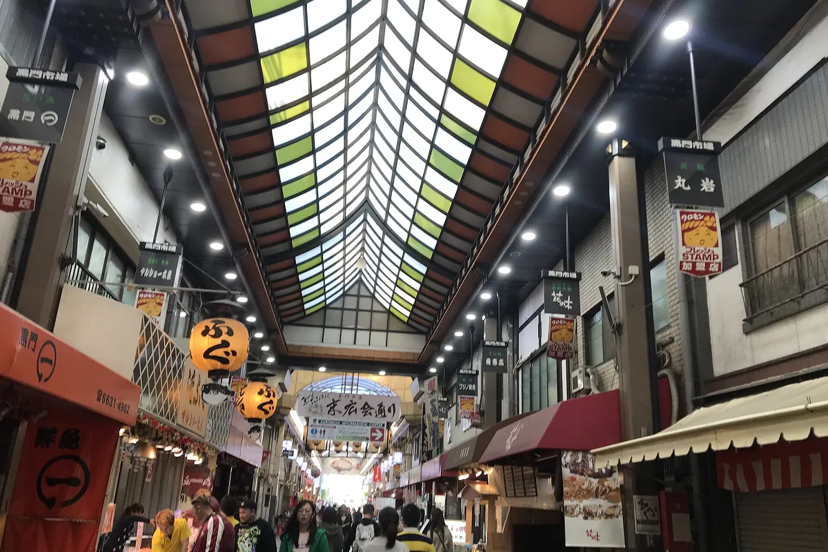 Shoppers walking through the roofed glass-ceilinged arcade of Kuromon Market in Osaka with shop signs