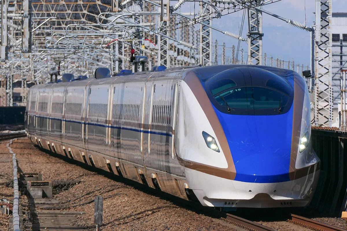 A Hokuriku Shinkansen E7 series bullet train running on tracks past overhead power line gantries