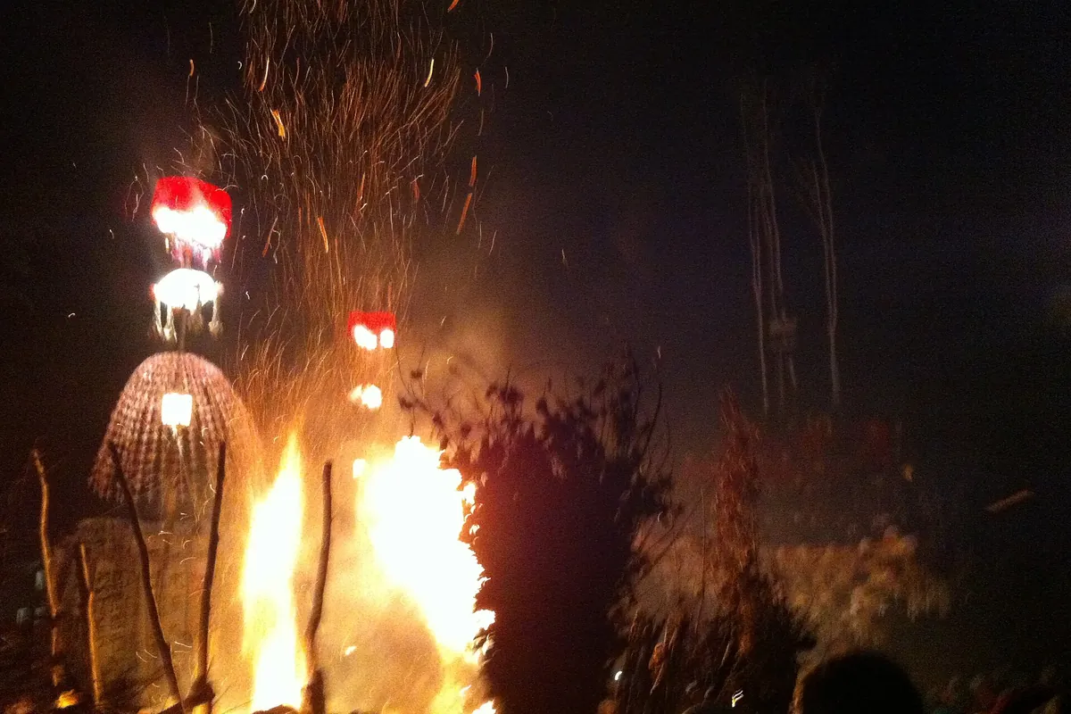 Burning wooden structure with flames and sparks at night, torches in the foreground, at the Dosojin fire festival in Nozawa O