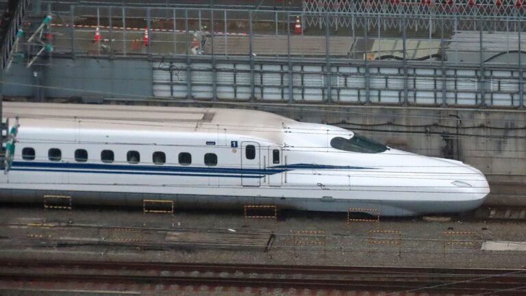 A white N700S Shinkansen train with a blue stripe running along elevated tracks beside safety fencing