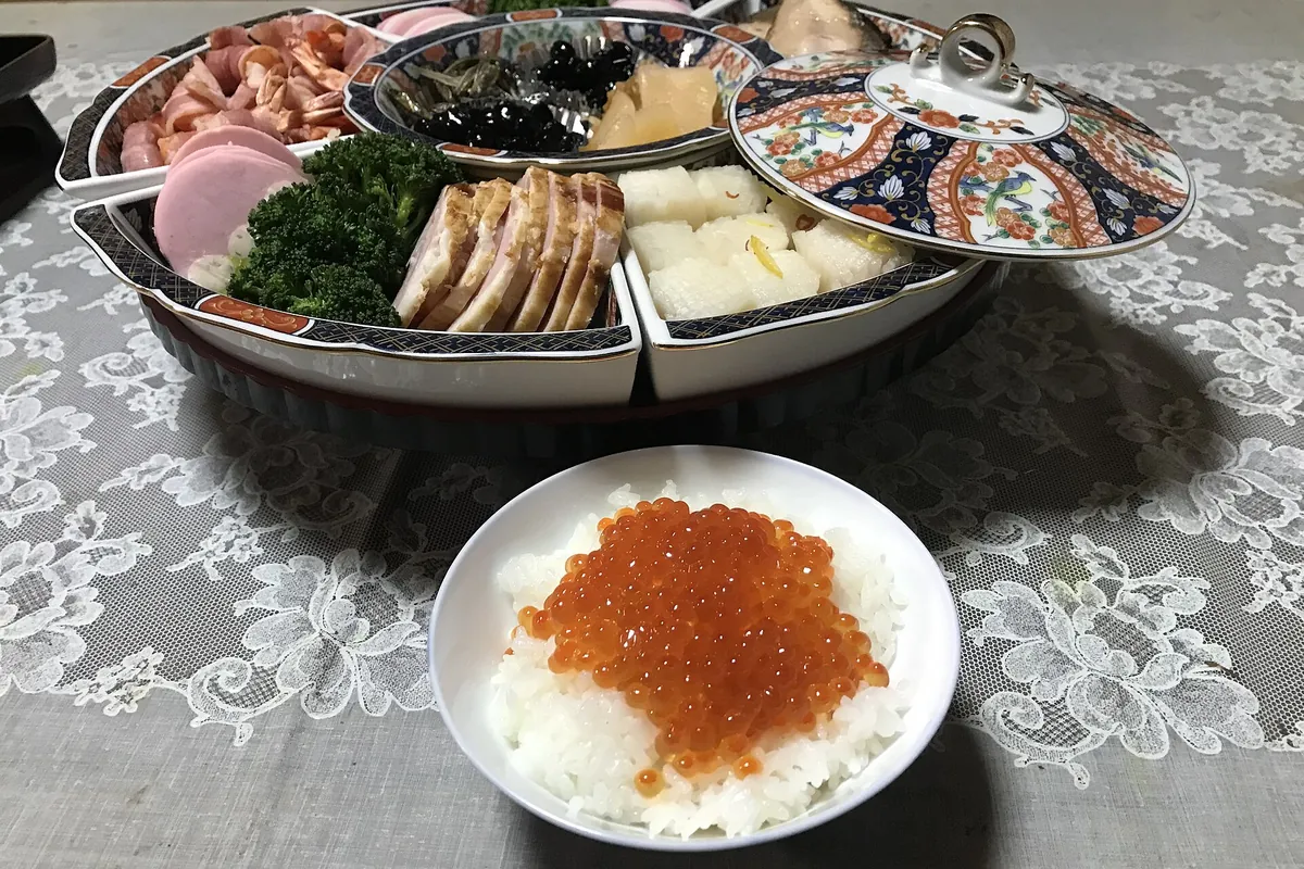A compartmented osechi tray with ham, broccoli, black beans, kamaboko and roast pork, beside a bowl of salmon roe over rice