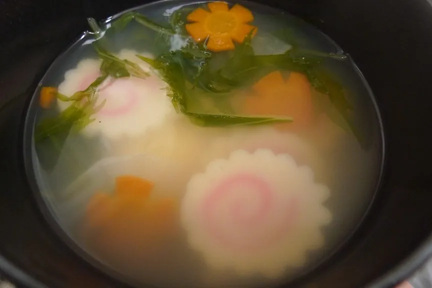 A bowl of ozoni soup with pink and white mochi, a flower-shaped carrot slice and greens in clear broth