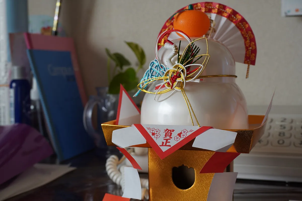 A kagami mochi New Year decoration of two stacked white rice cakes, an orange, red paper fan and cords on a gold stand